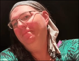 Close-up portrait of a person wearing glasses, a gray patterned headscarf, and a green patterned top, looking slightly up toward the camera. The background is dark.