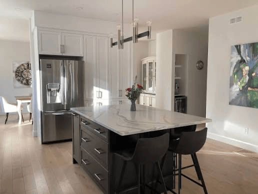 Bright modern kitchen with light wood floors, white cabinets, and a stainless steel refrigerator. A large dark island with a white marble top holds a vase of flowers and has two black stools beneath it, with a contemporary light fixture above and a colorful painting on the wall nearby.