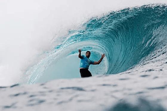 A surfer in a blue shirt rides inside a large curling turquoise wave, with arms raised for balance. The foamy water fills the foreground and the wave arches overhead.