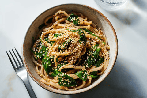 A bowl of thick noodles topped with wilted greens, sesame seeds, and a light sauce sits on a white table, with a fork at the left and a glass partially visible at the top right.