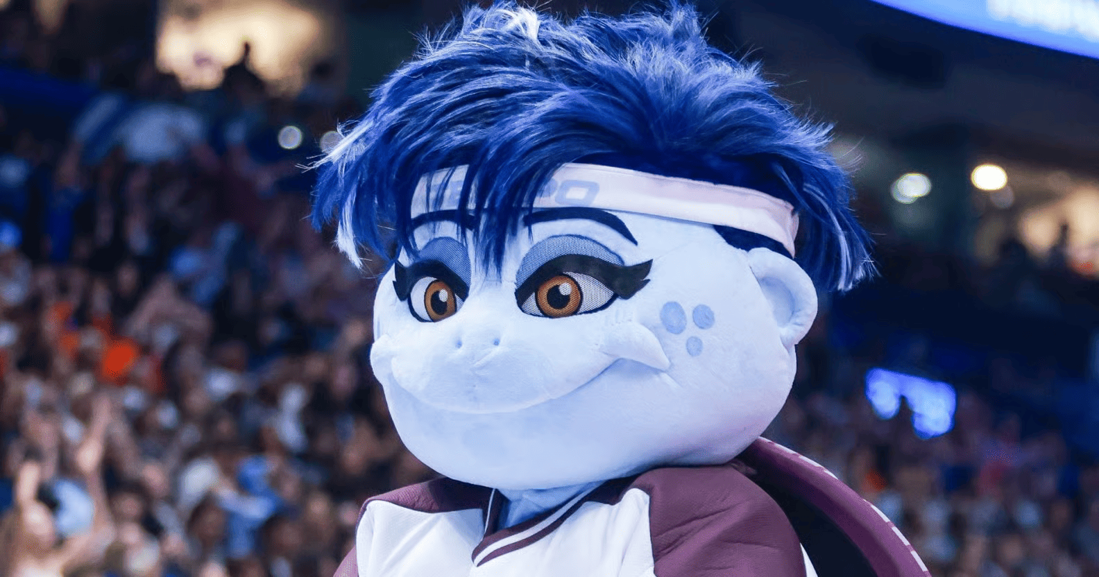 A close-up of a smiling blue-and-white mascot with spiky blue hair, a white headband, large brown eyes, and a maroon-and-white jersey. A blurred crowd and arena lights fill the background.
