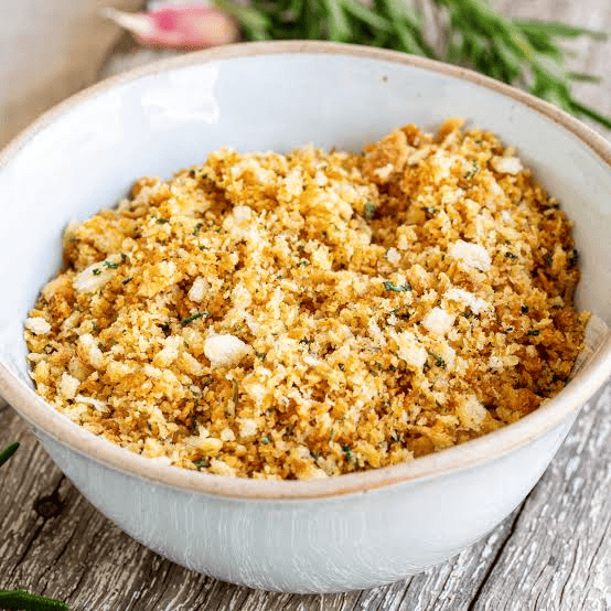 A white bowl filled with golden seasoned breadcrumbs or stuffing crumbs, speckled with herbs, sits on a weathered wooden surface. Green herbs and a garlic clove are softly blurred in the background.