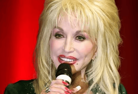 A blonde woman with voluminous hair sings into a microphone against a bright red background, wearing sparkling clothing and heavy makeup.