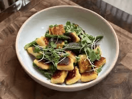 A shallow bowl holds roasted golden-brown gnocchi pieces topped with microgreens and a light drizzle of sauce. The dish sits on a wooden tabletop.