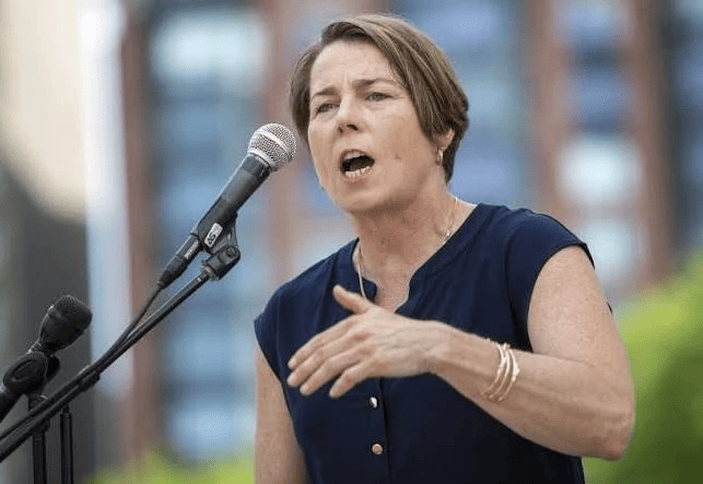 A woman with short hair speaks emphatically into a microphone at an outdoor event, gesturing with one hand while wearing a navy sleeveless top and gold bracelets. The background is blurred with buildings and greenery.