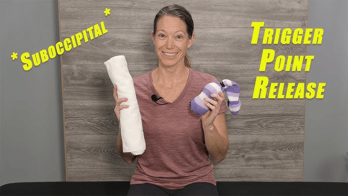 A smiling woman sits in front of a wood-grain backdrop holding a rolled white towel and a purple striped sock, demonstrating a neck exercise. Large yellow text reads “* SUBOCCIPITAL * TRIGGER POINT RELEASE.”