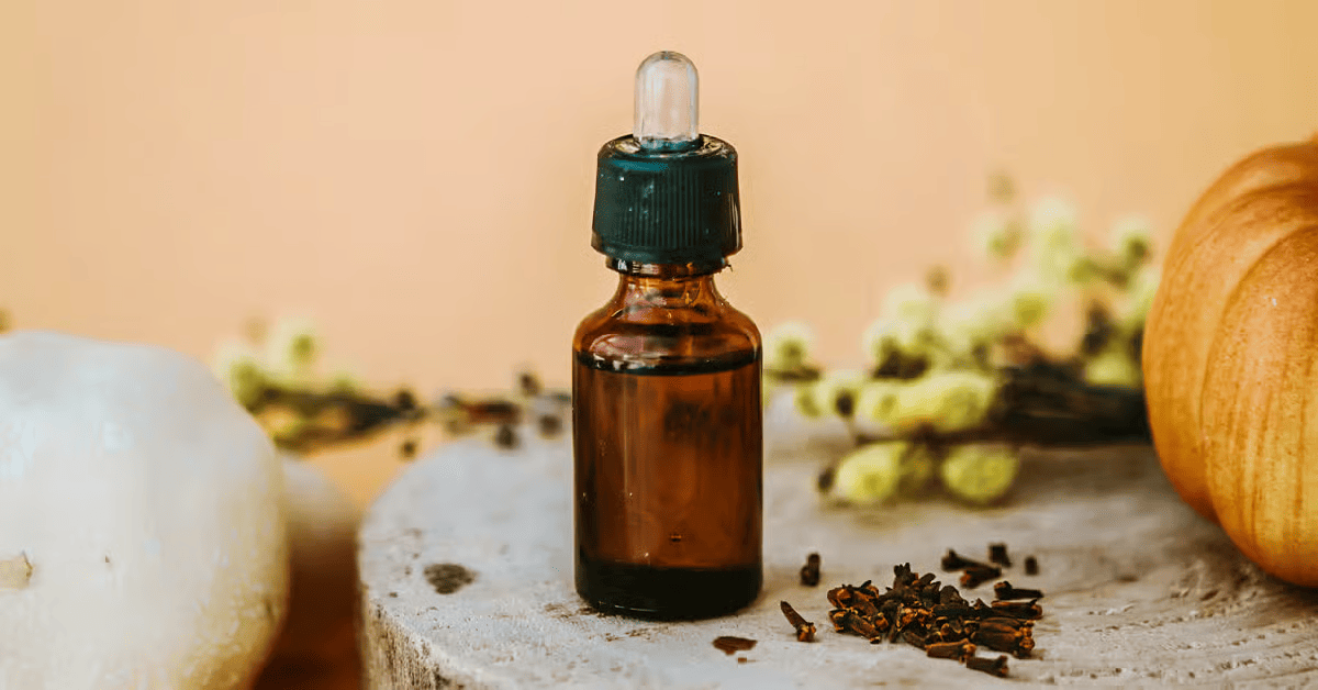 An amber glass dropper bottle of clove oil stands on a rustic surface surrounded by pumpkins, cloves, and pale flowers against a warm beige background.
