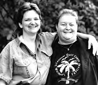 Brenda and Wanda Henson: Two smiling women stand arm in arm in front of leafy foliage, with one wearing a T-shirt printed with a circular palm-tree logo and the text “3rd Annual Gulf Coast Women’s Festival” and “1991 Biloxi.”