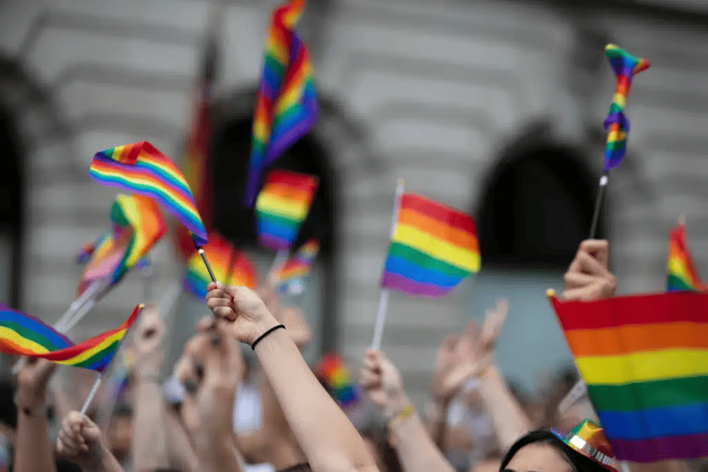 A crowd raises rainbow Pride flags in front of a large building, with many hands and flagpoles visible against a blurred background.