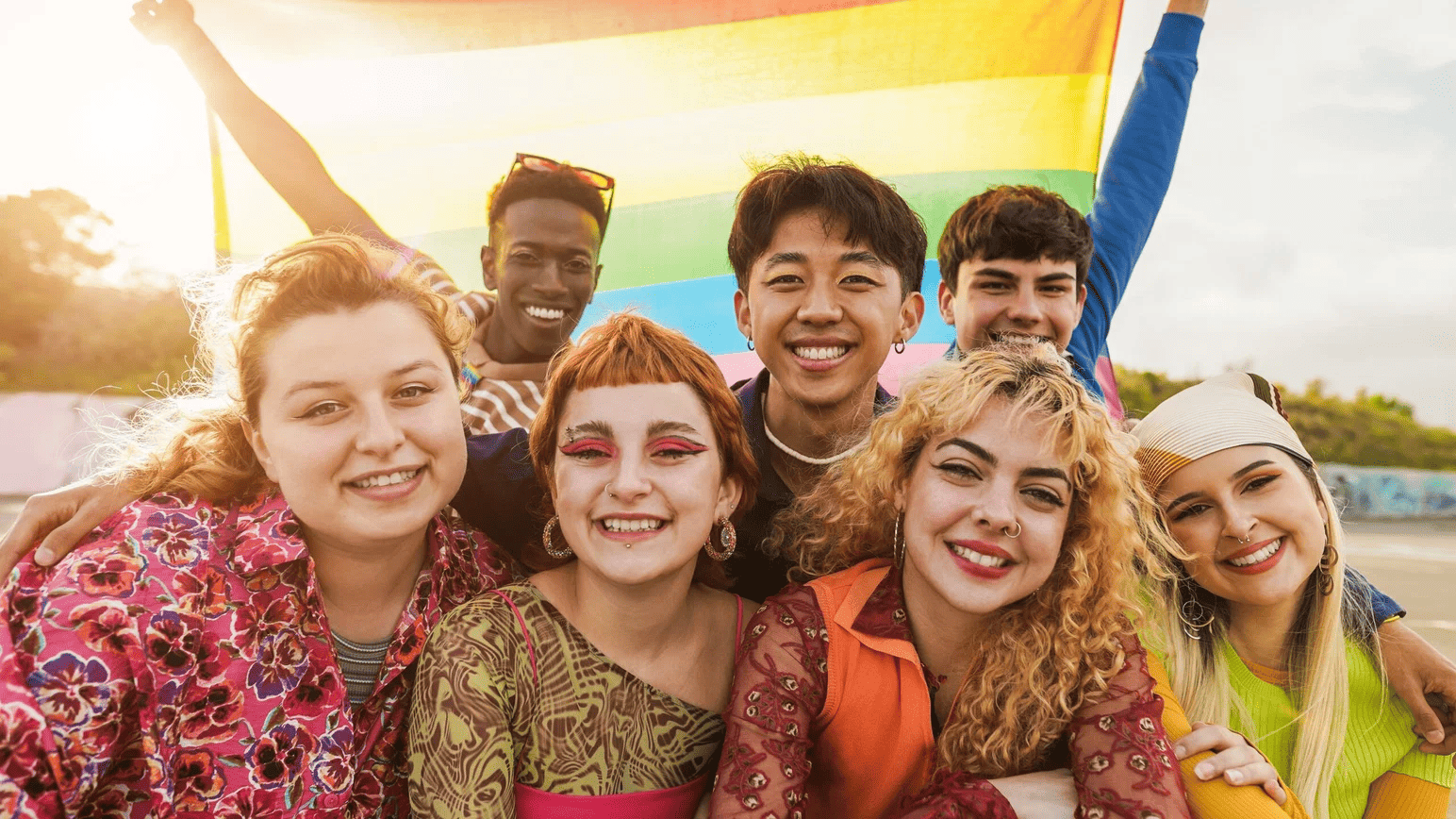 Six smiling young adults pose closely together outdoors in front of a large rainbow pride flag, with two people raising their arms in the background. The group includes diverse hairstyles, clothing, and expressions, and the scene is brightly backlit by the sun.