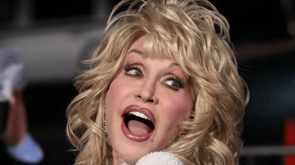 A blonde woman with curled hair and heavy makeup is caught mid-performance, mouth open wide and eyes looking upward. The background is dark and out of focus.