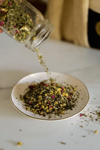 A glass jar is tipping dried herbs and flower petals onto a white plate, forming a small pile on a light tabletop with a few pieces scattered around.