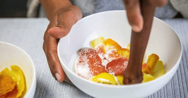 Hands hold a white bowl of sliced fruit sprinkled with white powder while a wooden pestle stirs or crushes the mixture. A second bowl with more fruit is partially visible on the left.