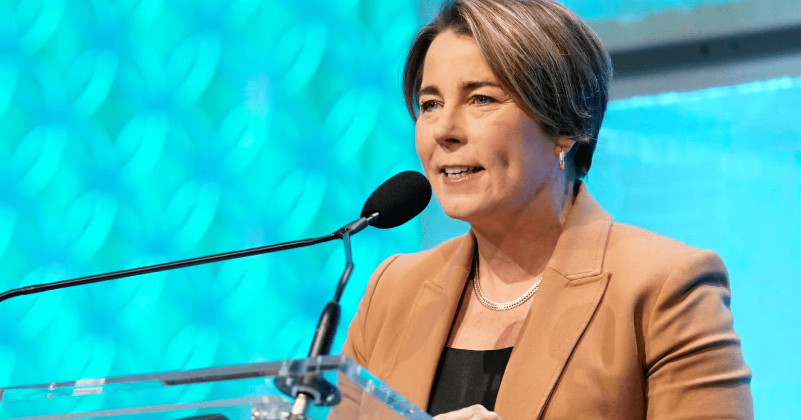 A woman in a tan blazer speaks at a clear podium with a microphone, against a bright blue patterned background, MA Governor.