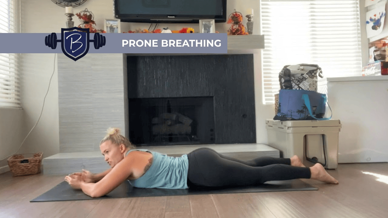 A woman lies face down on a yoga mat in a living room, propped on her forearms and facing left, with a fireplace behind her. A banner across the top reads “PRONE BREATHING” with a stylized “B” logo.